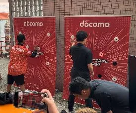 docomo ブランドの REAXION チャレンジパネル前で対戦する 2 人とそれを撮影する観客